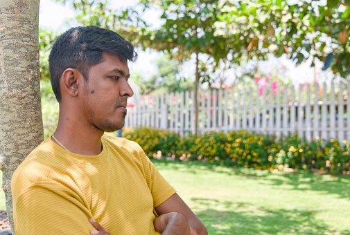 An Indian man sits on a bench in a peaceful garden, lost in thought on a bright sunny day.