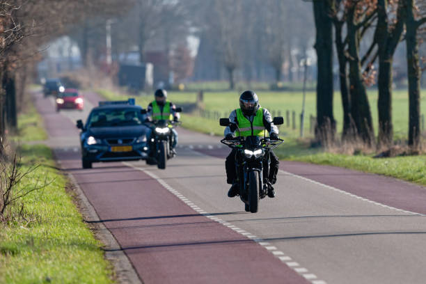 The Netherlands, Overijssel, Twente, Enschede, Lonneker, February 7th 2023, daytime front view close-up of two caucasian male bikers approaching on an asphalt road with bicycle lanes on both sides in a rural setting, taking motorbike training lessons followed by the instructor driving in a car, giving them instructions by means of a headset