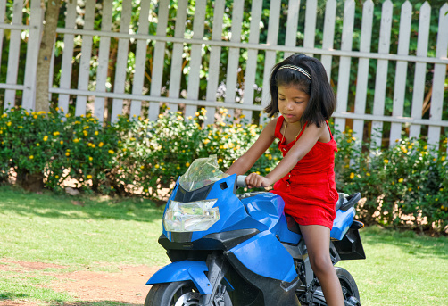 A joyful young girl with bright eyes and a smile rides a small toy motorbike on a sunny day in a green backyard