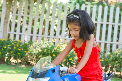 A young girl with bright eyes and a red dress enjoys a carefree moment riding a toy motorbike in a sunny garden.