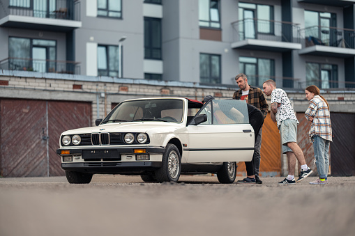 Riga, Latvia - April 1, 2024: A group of young people looking with interest into white classic convertible BMW E30 car. Old garages and a modern building in the background.