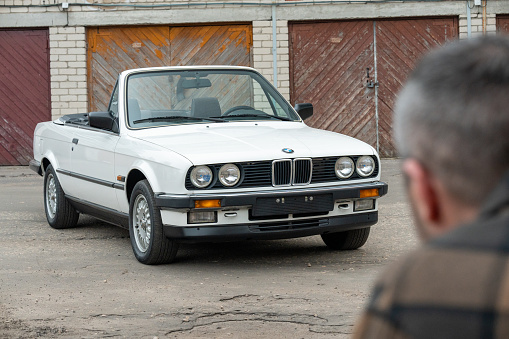Riga, Latvia - April 1, 2024: A man looking on his white classic convertible BMW E30 car with opened roof. Old garages in the background.