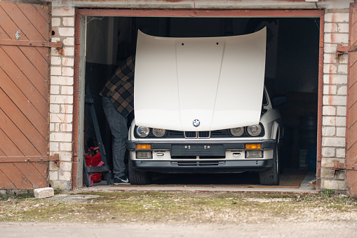 Riga, Latvia - April 1, 2024: A man under the hood of white classic convertible BMW E30 car in the opened garage.