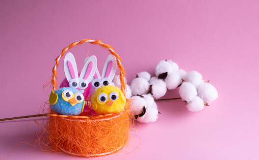 A branch of white cotton, toy eggs with birds in an orange basket and Easter bunnies on a pink background