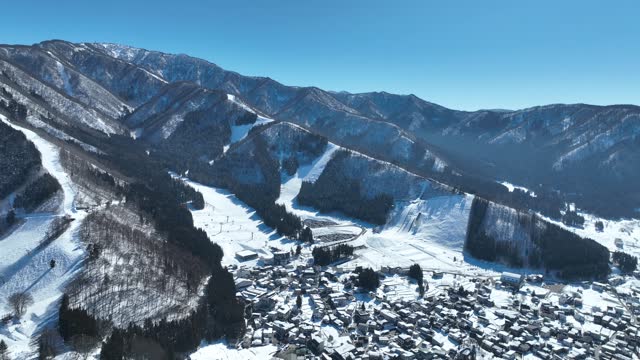 Aerial establishing shot of Japans Nozawaonsen Mountain Ski Resort Village. Camera slowly descends and pans up revealing the mountains and ski fields. Clear Winter Day