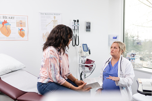 The expecting female physician gestures while helping her patient understand the changes to her medication regimen.