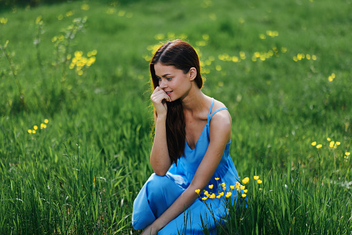 Woman in spring in nature smile with teeth sitting in a field on the green grass with flowers and smiling happy sunset in a blue dress with red hair. High quality photo