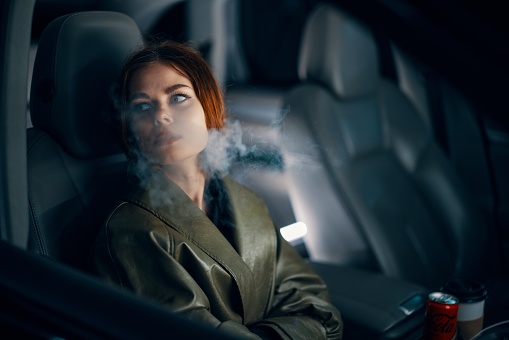 a close horizontal portrait of a stylish, luxurious woman in a leather coat sitting in a black car at night in the passenger seat, looking to the side through the fogged glass. High quality photo