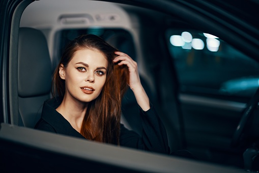 a stylish, luxurious woman sits in a black car at night in the passenger seat, and looks pleasantly into the camera. Close horizontal photo. High quality photo
