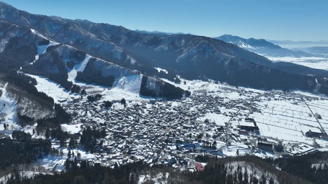 Aerial orbiting shot of Japans Nozawaonsen Mountain Ski Resort Village. Camera orbits the ski village resort with mountains and ski fields in background. Clear Winter Day