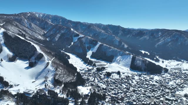 Aerial establishing shot of Japans Nozawaonsen Mountain Ski Resort Village. Camera orbits the ski village resort with the mountains and ski fields in background. Clear Winter Day