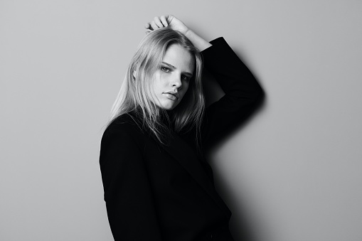 Serious young blonde woman leans on the studio wall holding hand overhead looks at camera posing isolated in black jacket. High quality photo