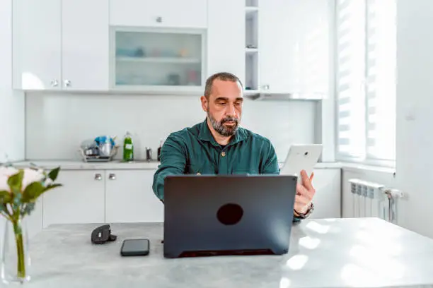 A mature man maximizes efficiency from his home office by expertly wielding both a laptop and a digital tablet, embodying the multitasking prowess required to thrive in today's fast-paced professional environment