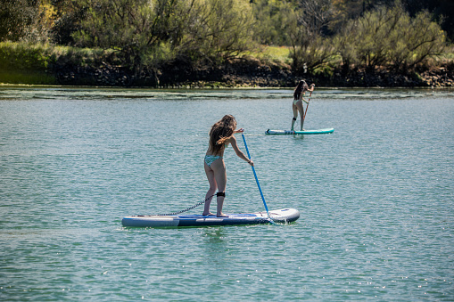 multicultural pair of bikini-clad women seen from behind as they paddleboard together on a sunny summer day on a tranquil lake, enjoying outdoor water sports and leisure activities