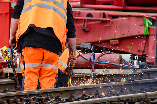a worker with an orange warning vest works with a sparking welding torch on a railroad track