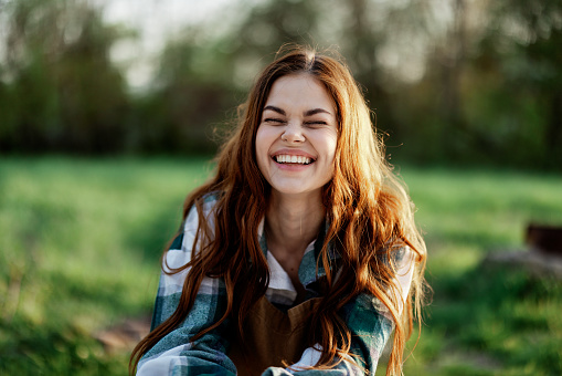 Funny woman portrait close-up laughing and wiggling for the camera in front of a green park in summer, the sunset shining on her red hair. High quality photo