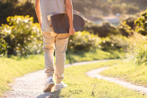Legs of a man walking outdoors on a path, carrying his longboard to find a spot to ride. Background showcases a natural setting with abundant trees