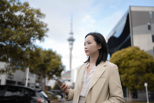 Young Asian woman in suit holding smartphone standing on city street with landmark Sky Tower in the background