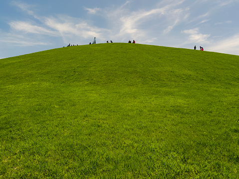 Tall green hill covered in lush green grass against a cloudy blue sky. Silhouettes of unrecognizable people gathered at the top of the hill for a viewing party.