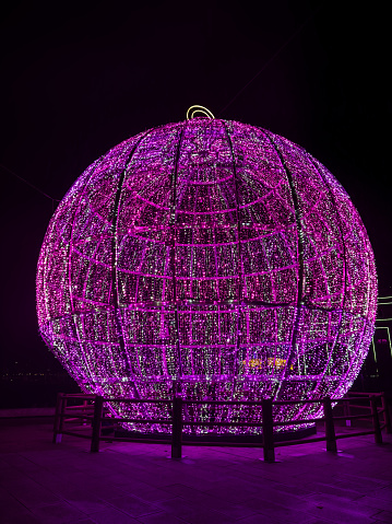 Amidst the quiet of night, a captivating orb of purple lights emerges as a luminous landmark against the dark canvas of the sky. This photograph captures the magical essence of the holiday season through a magnificent display of lights, arranged meticulously to form a gigantic glowing sphere. This radiant structure, a modern interpretation of a festive bauble, illuminates the surrounding area with a warm, inviting glow, symbolizing hope and joy. The lights reflect the spirit of the season, inviting passersby to pause and bask in the enchanting atmosphere created by this urban installation.