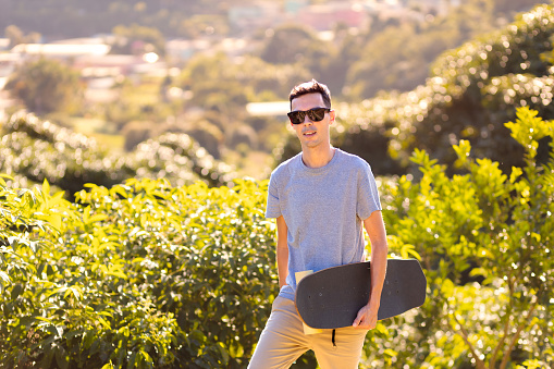 Man walks outdoors, carrying his longboard, seeking a spot to ride. Background showcases a natural setting with abundant trees. Promotes outdoor activities and exploration