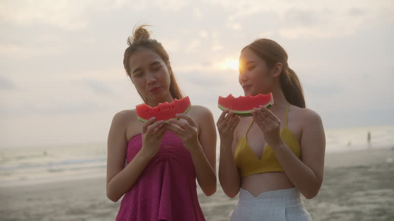 Two beautiful young friends having fun eating watermelon and coconut on the beach at sunset.