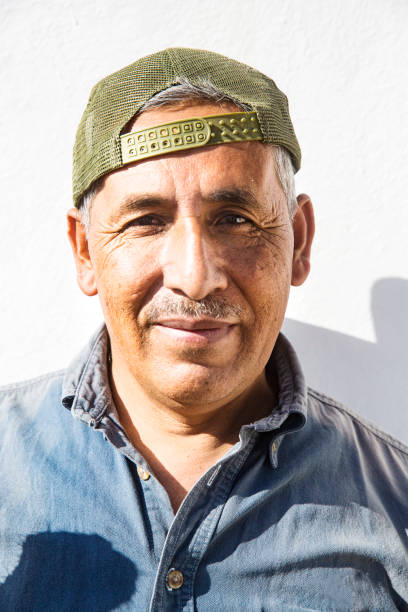 A middle aged man with a strong face, wearing a backwards baseball cap, against a white background lit by strong sunlight