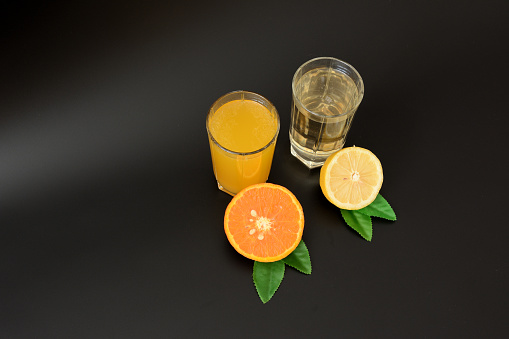 Two tall glass glasses with citrus juices on a black background, next to the halves of ripe orange and lemon with leaves. Close-up.