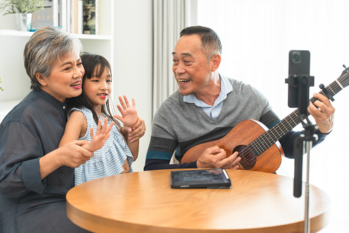 Asian family, grandfather playing guitar for grandmother, grandchild sitting in living room at home, singing and having fun together, recording video, online streaming on social media via mobile phone