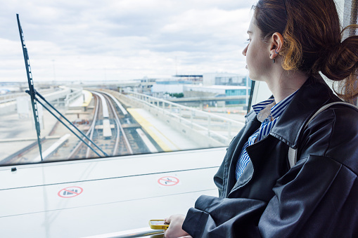 Young Caucasian businesswoman on a business trip, standing at an airport station and looking at her phone. The background shows rail tracks and a part of the rail station structure at John F. Kennedy International Airport