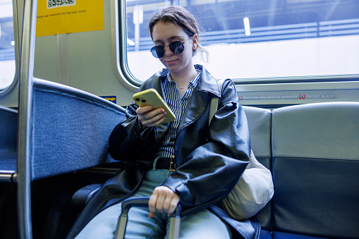 Young Caucasian businesswoman, in sunglasses, sitting on a train and engaging with her smartphone. Woman managing work-related communications and organizing her schedule