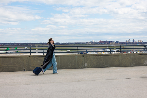 Young Caucasian woman, dressed in a coat, walks alongside an airport with a suitcase. She is on a business trip, hanging on smartphone. Airplane taking off in the background, JFK airport runways and the city skyline in the distance.