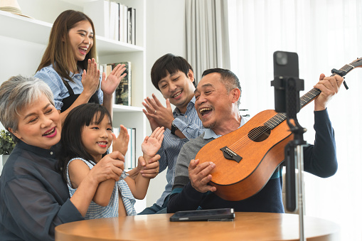 Happy Asian three generation family, grandparent, mother, father, daughter playing guitar, singing and having fun together, sitting in living room at home, recording video with mobile phone