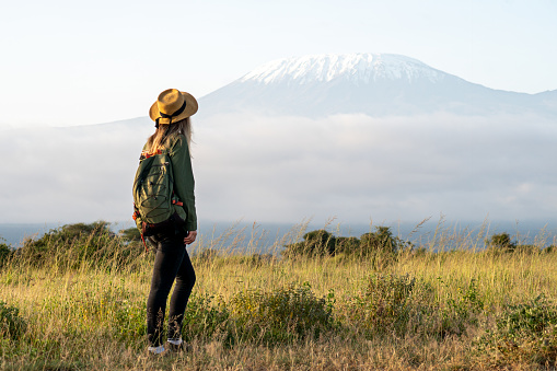 girl walks against backdrop of Kilimanjaro. African walking safari. woman traveler with backpack looking at amazing mountains and forest, wanderlust travel concept, copy space, atmospheric epic moment