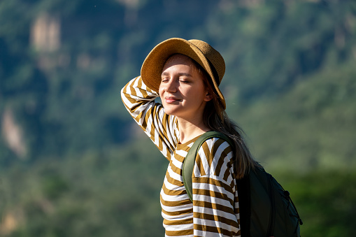 smiling woman in the hat with backpack travels through the mountains and rests on the top of a mountain on a summer sunny day. Attractive girl enjoying healthy lifestyle outdoors
