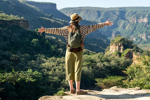 Rear view of an enthusiastic woman in a hat with a backpack with her hands raised, looking from height at an amazingly beautiful landscape of mountains. Concept of freedom and adventure.