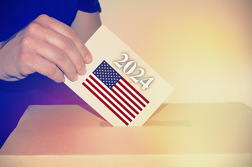 Hand in polling station, voting in a booth with US flag in background.