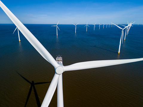 A wind farm in the middle of the ocean in the Netherlands Flevoland, with windmill turbines gracefully spinning under the spring sky. drone aerial view of windmill turbines green energy in the ocean