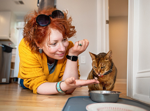 Redhead woman feeds her cat from her hand, which the cat licks off her fingers. The woman smiles and offers food to her cat, lying on the floor in the kitchen near her pet's bowl