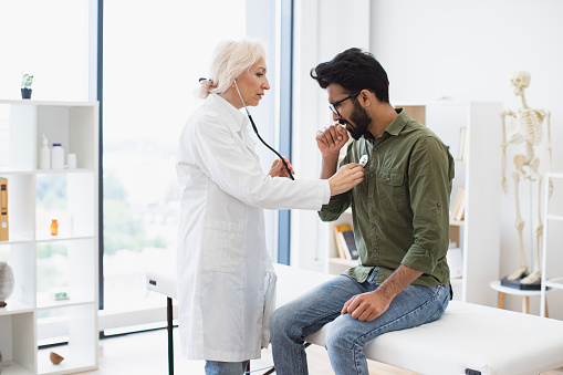 Elderly woman in white lab coat checking lungs and heart via stethoscope. Serious bearded man in everyday wear visiting family doctor for full examination in general practice.