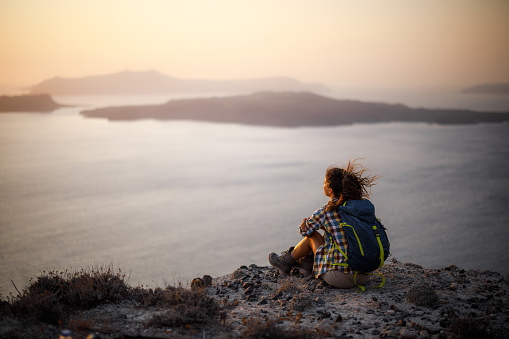 Back view of a female backpacker taking a break after hiking on a hill above the sea at sunset. Copy space.