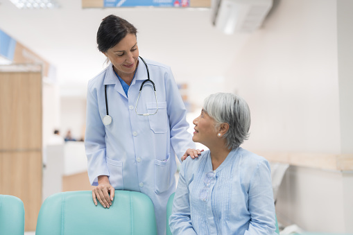 A serious female doctor gestures as she discusses healthcare options with a mature female patient. The patient is reading an informational brochure.