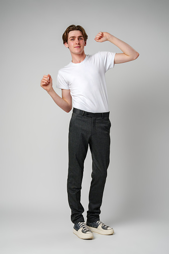 A cheerful young man with a playful expression is caught mid-dance against a plain background, wearing a simple white T-shirt, gray pants, and white sneakers. His dynamic posture suggests a sense of rhythm and carefree enjoyment.