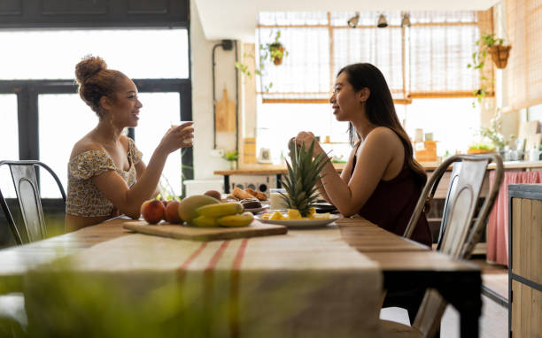 Two Friends Sharing Laughter over Breakfast A heartwarming scene of two young women sharing a joyful conversation during a healthy breakfast in a cozy kitchen. gay dinner table stock pictures, royalty-free photos & images