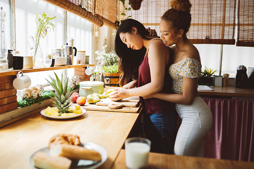Two young women bond while slicing fruit together, surrounded by a homey kitchen's morning glow.