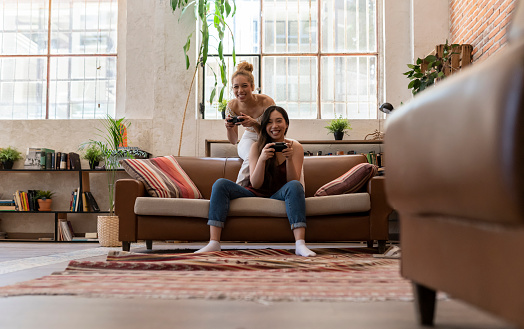 Laughter and rivalry light up the room as two friends enjoy a playful video game competition in a cozy, plant-filled living space.