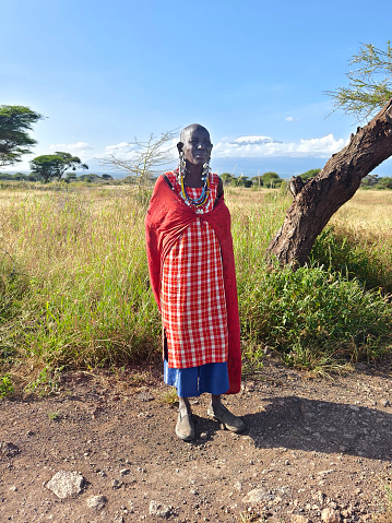 February 5, 2024. Amboseli National Park. Kenya: An adult Maasai woman in a traditional red dress and beaded jewelry stands against the background of the African savannah.
