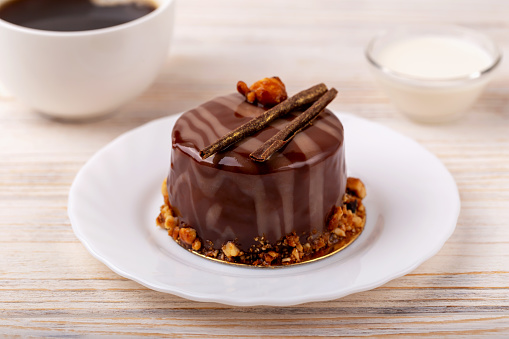 Fresh delicious chocolate cake on plate on white wooden background. Caramel glaze and decoration add appeal and desire. Cup of coffee and bowl of cream are nearby