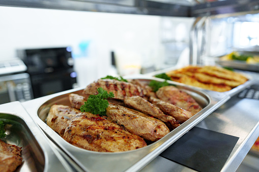 Grilled chicken breasts with appealing grill marks are garnished with parsley, paired with juicy pork cutlets, presented in a stainless steel serving tray as part of a buffet spread.