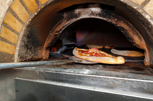 A pizza is placed inside a brick oven to bake. The heat from the oven cooks the pizza evenly, creating a crispy crust and melting cheese. The flames inside the oven dance around the pizza, giving it a smoky flavor.